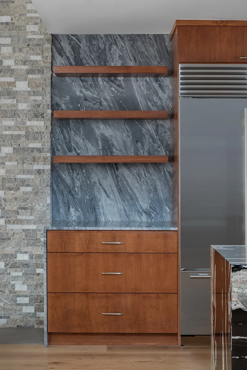 Kitchen cabinetry with gray marble backsplash, floating wooden shelves, and stainless steel refrigerator next to wood drawers.