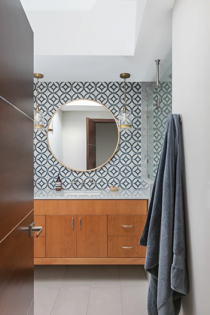 Bathroom vanity features a circular mirror and a geometric tile backsplash with hanging glass pendant lights.