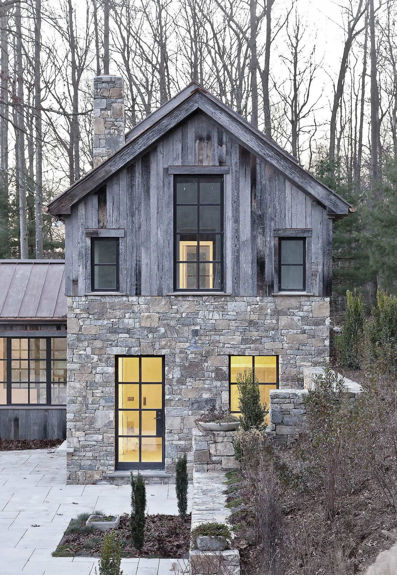 This stone and weathered wood exterior features large black window frames and a tall stone chimney in the Roxbury House project.