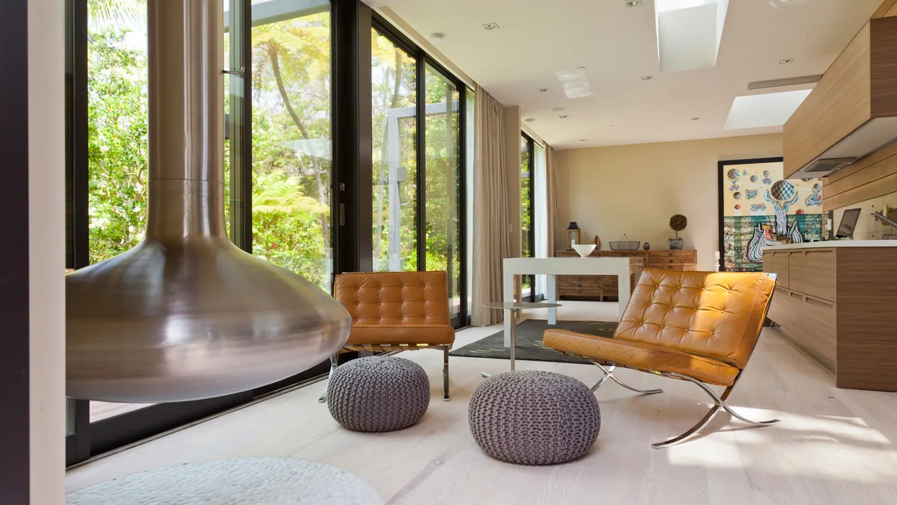 Living space featuring tan leather chairs, knitted poufs, a suspended silver fireplace, and large glass doors to a garden.