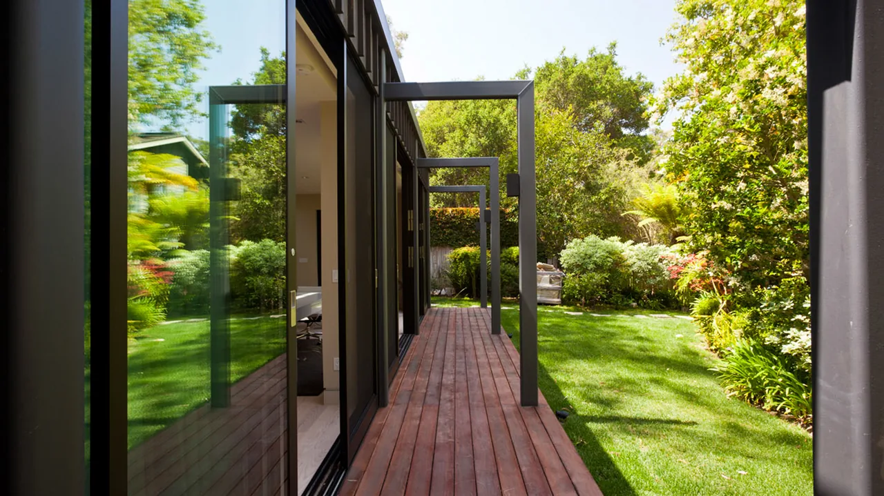 Exterior view of a wooden deck with steel pergola frames adjacent to glass sliding doors and a lush backyard.