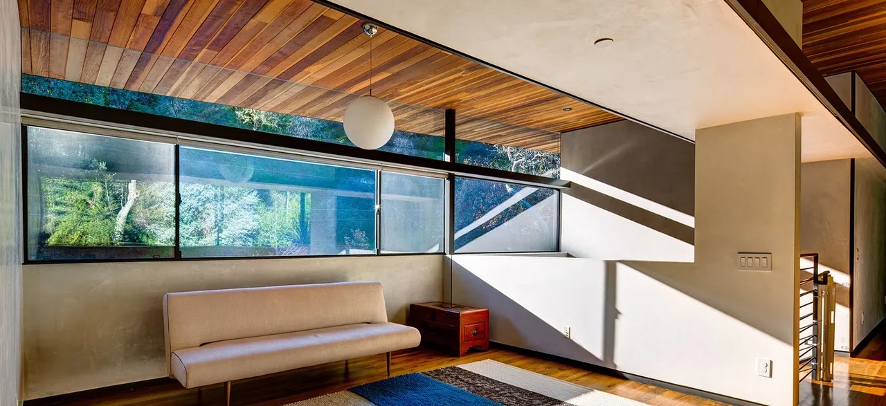 Living area features a wooden ceiling, large corner windows, and minimal furniture including a beige sofa and side table.