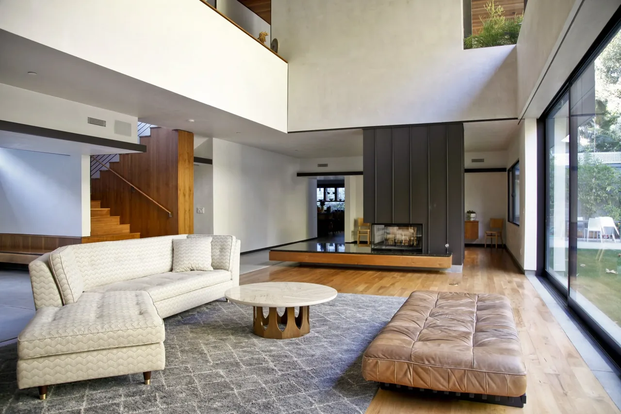 The Tree House living area by Jessie Bornstein features wood flooring, a central divider fireplace, and expansive glazing.