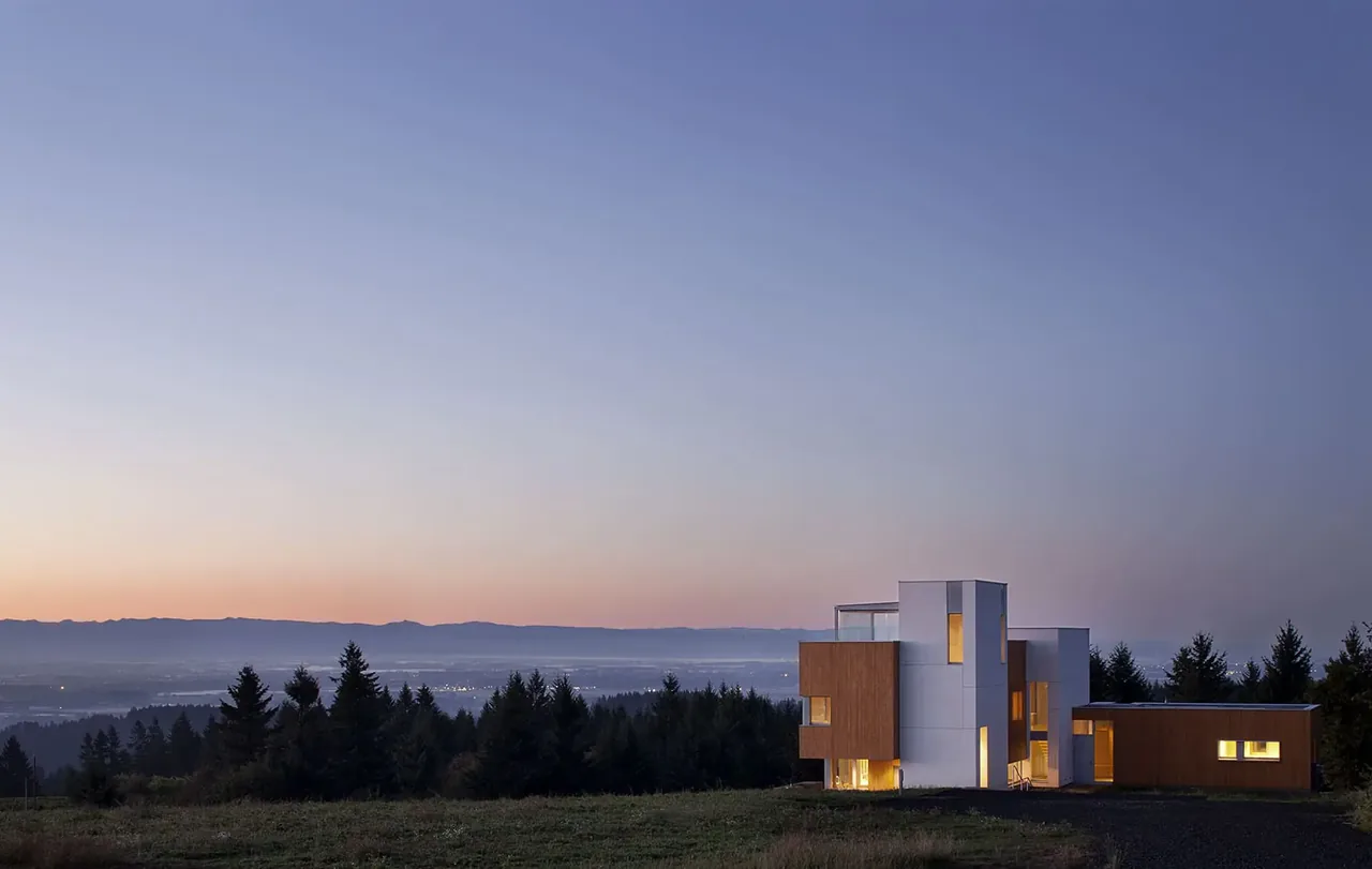 Karuna House by Holst Architecture P.C. sits on a grassy hilltop with forest and mountain views at sunset.