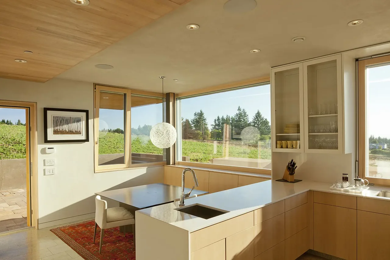 The Karuna House kitchen features light wood cabinets, a built-in bench dining nook, and expansive windows with wood frames.