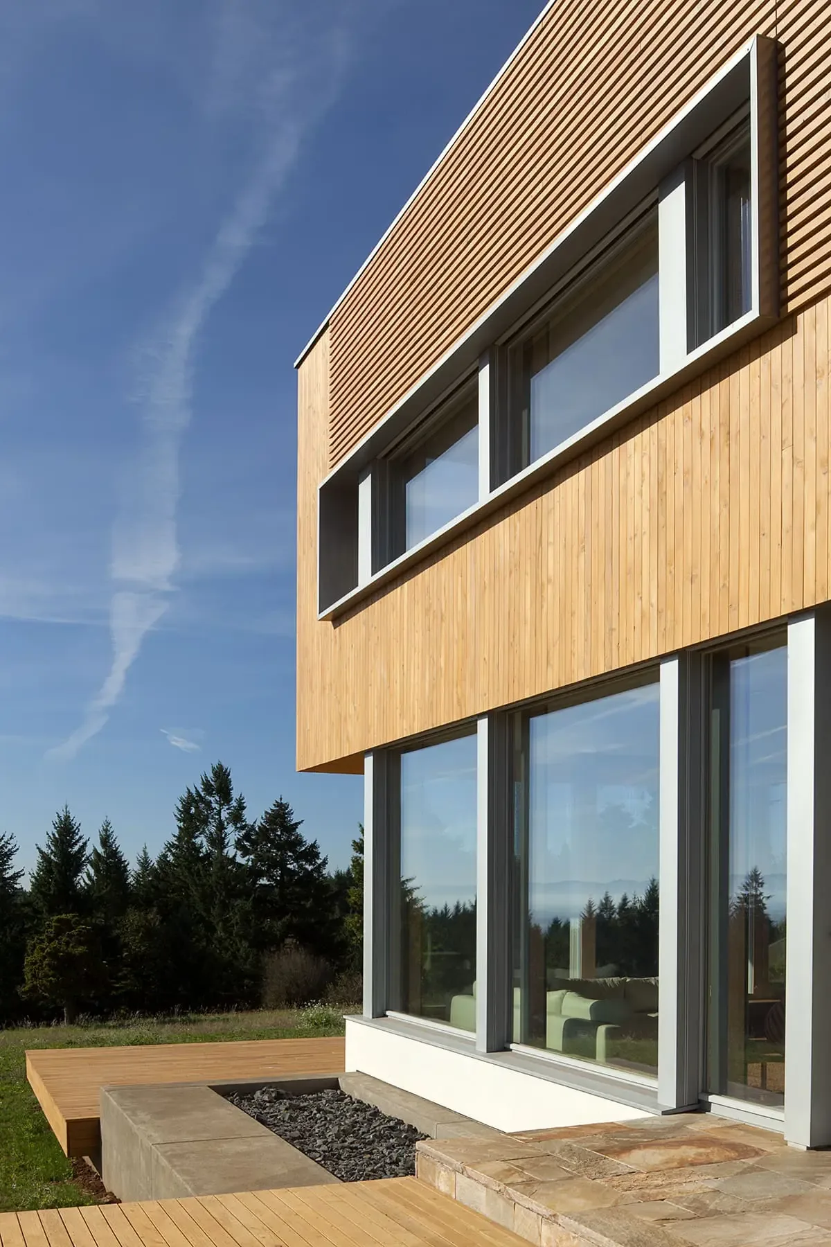 Karuna House exterior showcasing vertical wood siding with large window openings and a stone patio area.