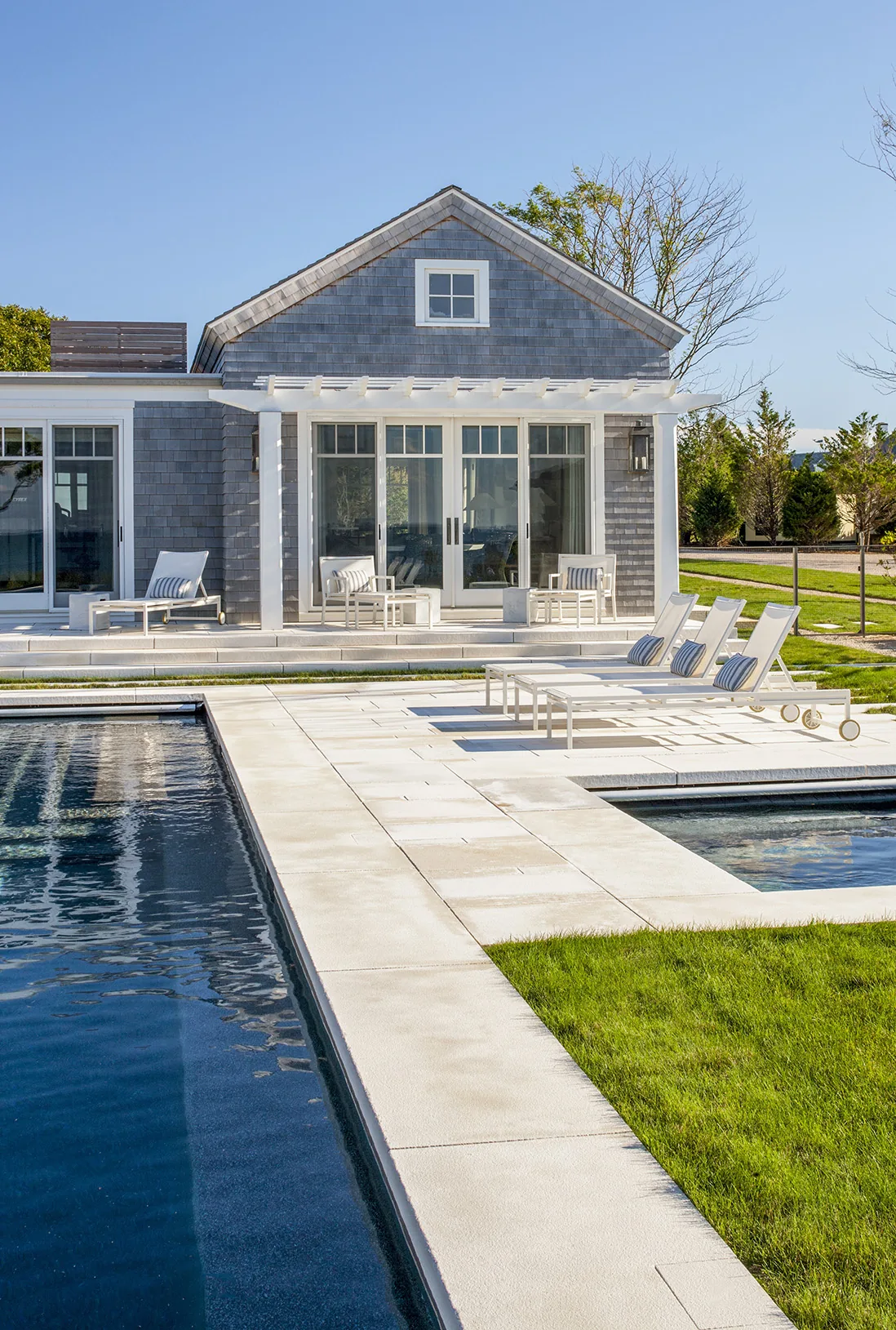 Backyard pool area with white stone deck, lounge chairs, and a gray shingle-sided house topped with a pergola.