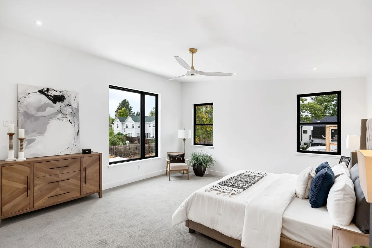 A minimalist bedroom features black window frames, a wooden dresser, and a ceiling fan with a light finish.