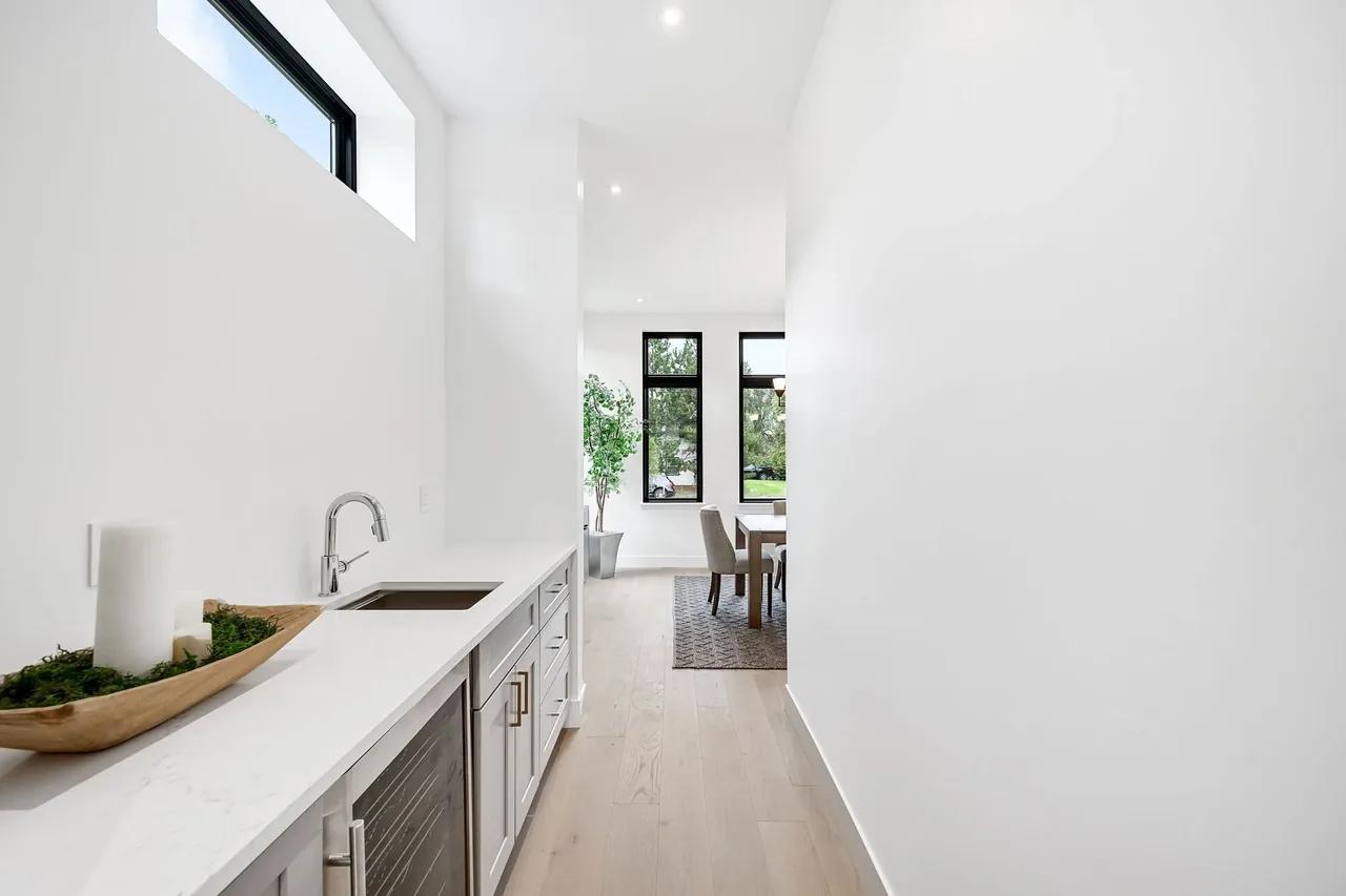 A bright, narrow kitchen hallway with a countertop and a small sink opens to a dining area with black-framed windows.