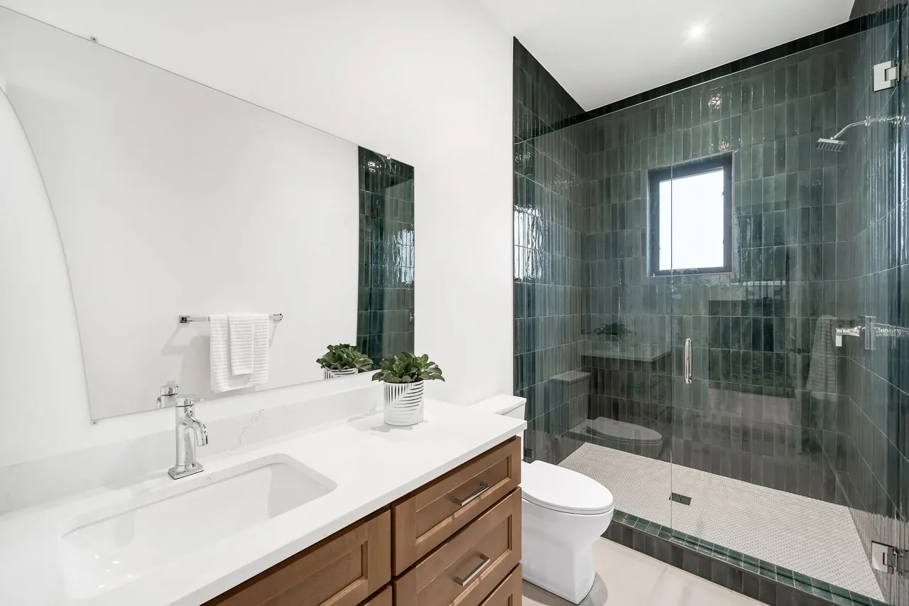Bathroom with a wooden vanity, white solid surface countertop, and a glass shower with dark green vertical tiles.