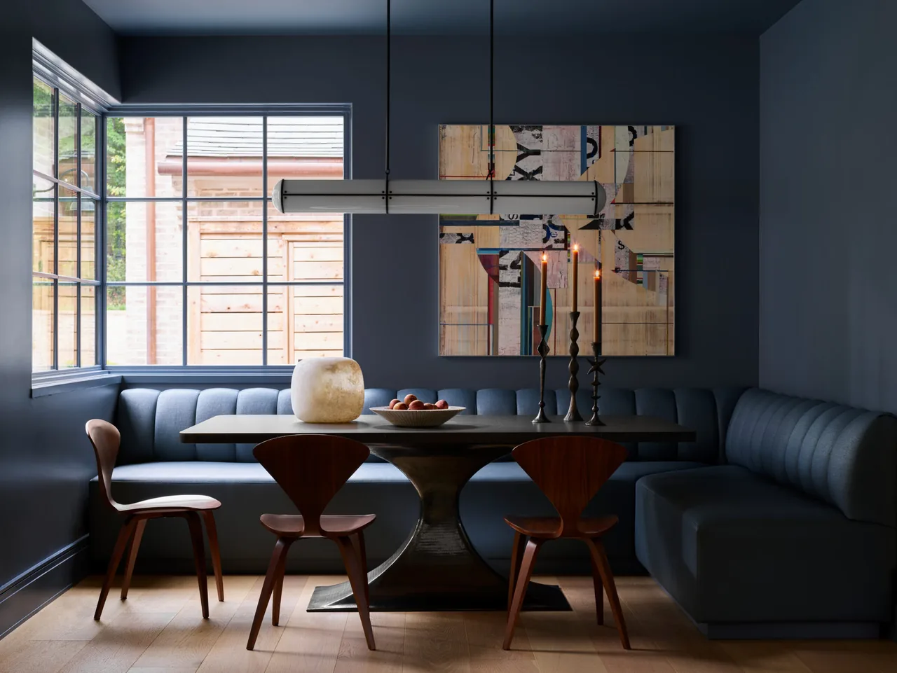 Dining nook with L-shaped banquette seating, wooden chairs, and a modern light fixture in a dark blue room.