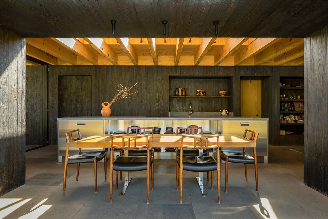 Dining area with wooden table seating six and a metal kitchen island in the background beneath exposed wood beam ceiling.
