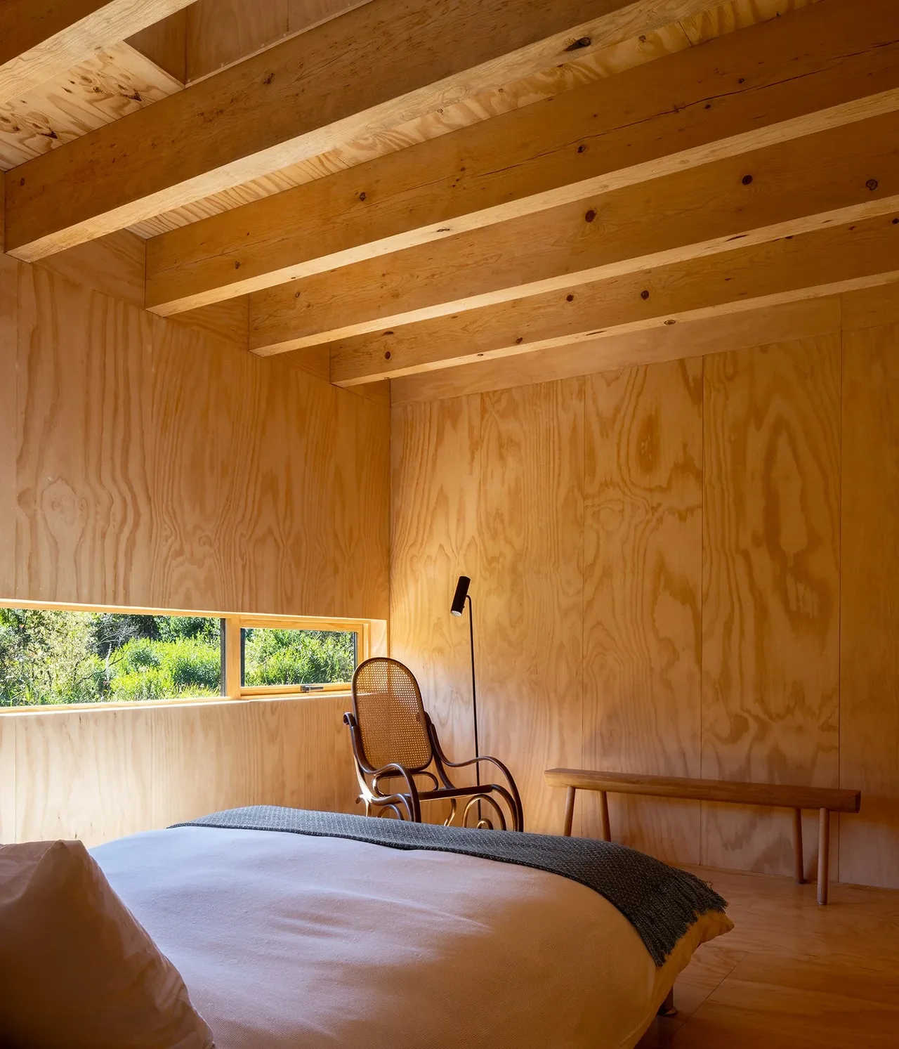 Bedroom features exposed wood beams, plywood walls, and a low horizontal window with views of natural landscape.