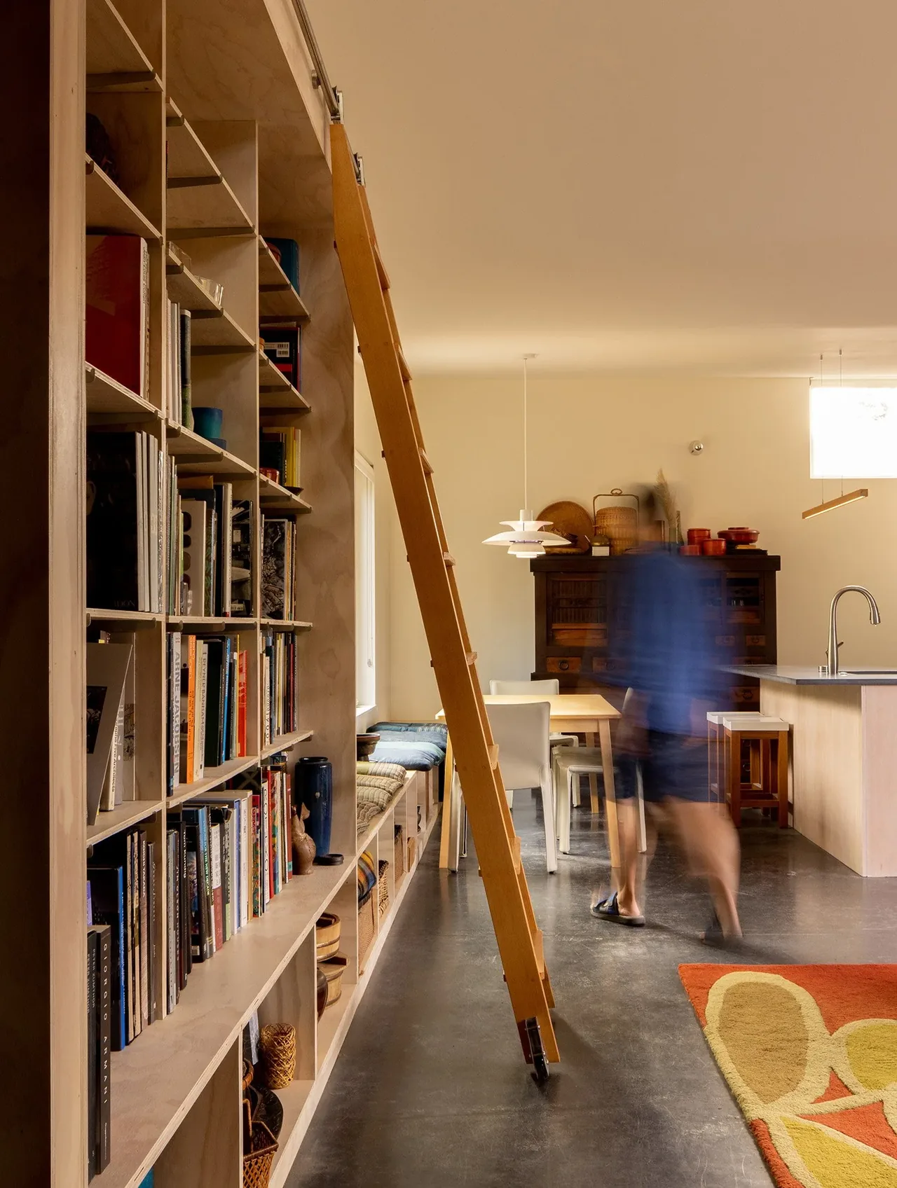 Room with built-in wooden shelving and rolling ladder, dining area, and kitchen island from House O by Hutchison & Maul Architecture.