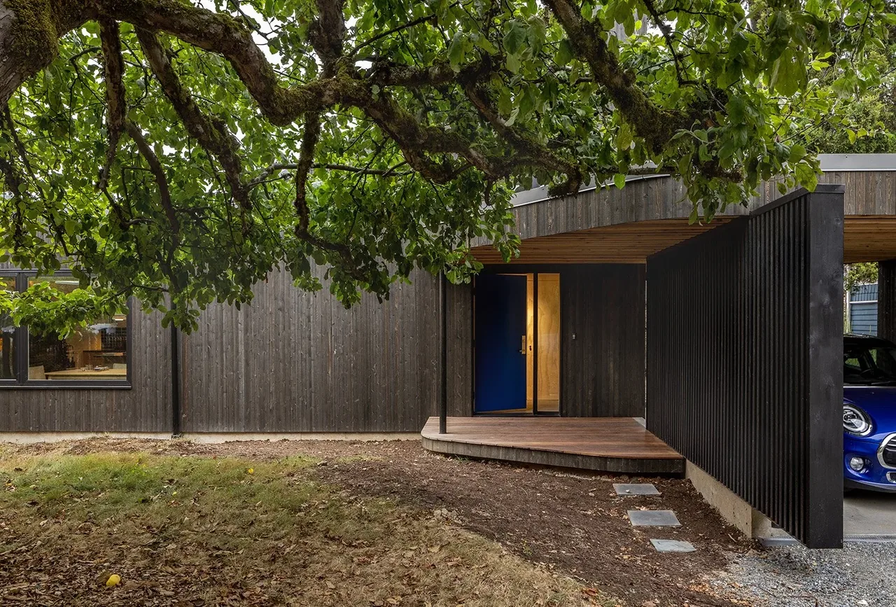 House O features dark wood siding, a blue entrance door, and a covered porch with a wooden deck, partially visible garage to the right.