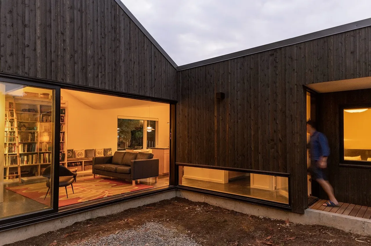 House O features dark wood siding with large windows revealing a cozy living room interior and a covered entryway.