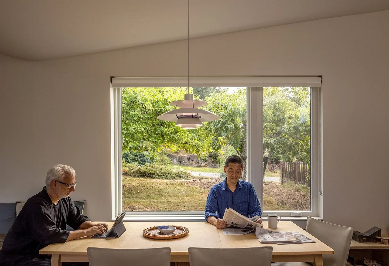 Interior dining area with a wood table faces a large window overlooking a garden.