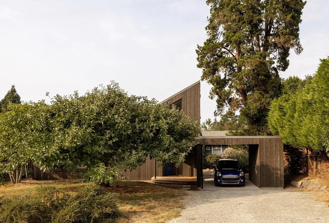House O features a wood exterior and a carport with a blue car parked inside, surrounded by mature trees and landscaping.