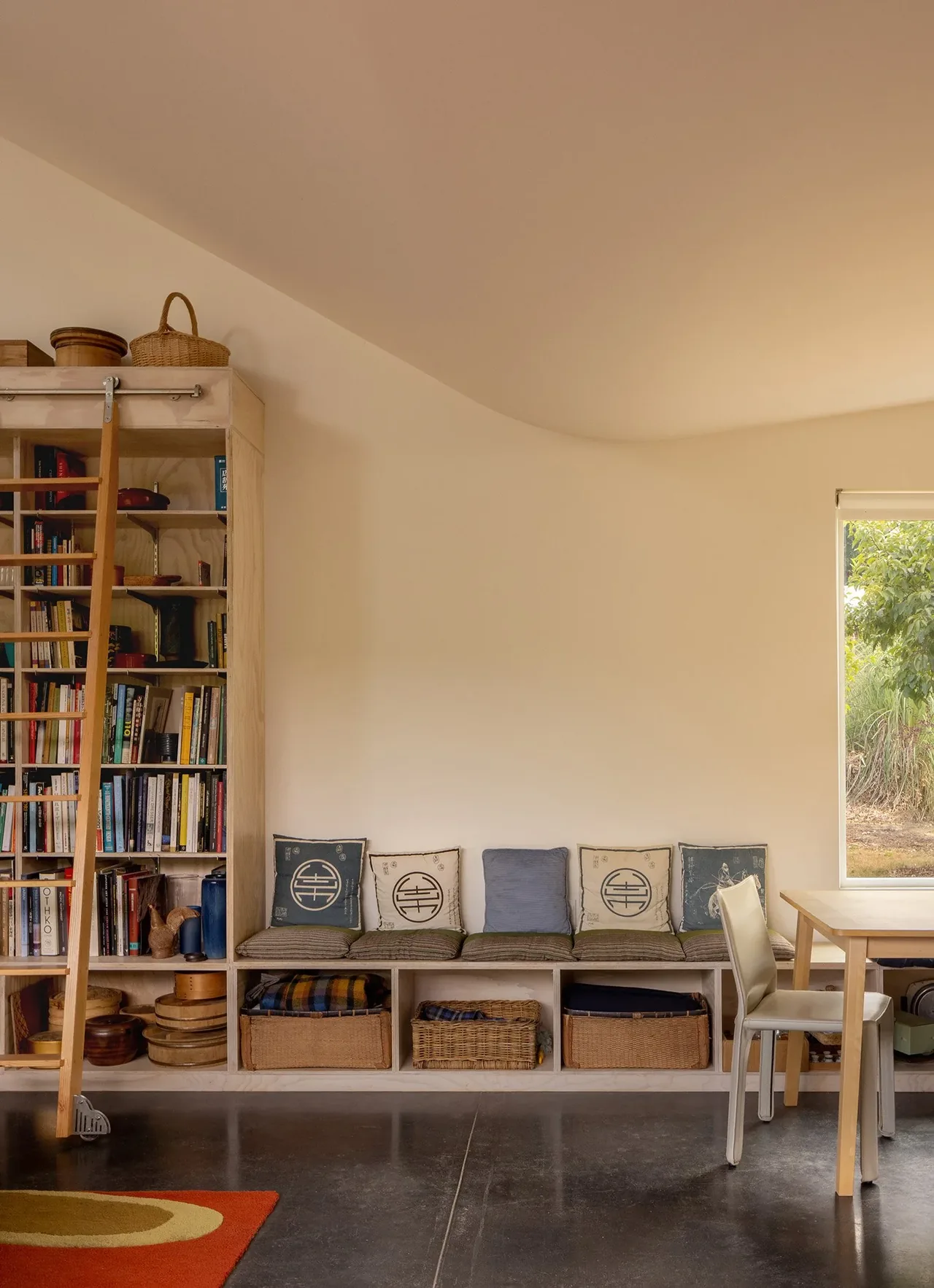 Built-in bench seating with cushions and pillows along a wall adjacent to a tall wooden bookshelf and a window looking outside.