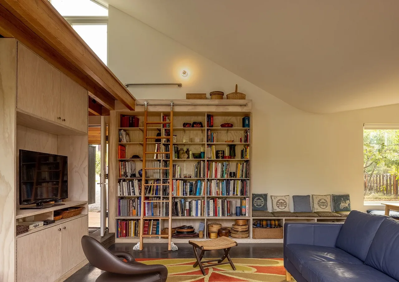 Living room of House O with built-in bookshelves, wooden ladder, and a blue couch near a large window.