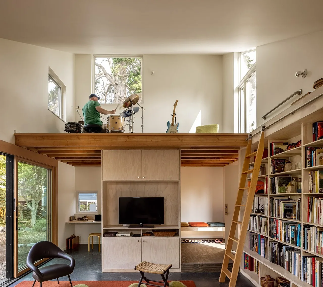 Living room with lofted music space, built-in shelving, and large windows revealing a green backyard view in House O.