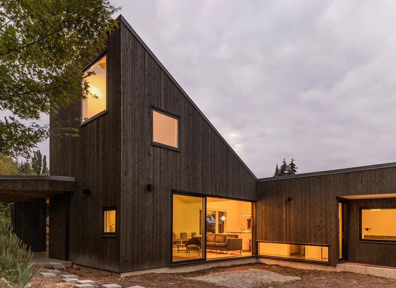 House O features dark vertical wood siding and large windows illuminating the interior living space at dusk.