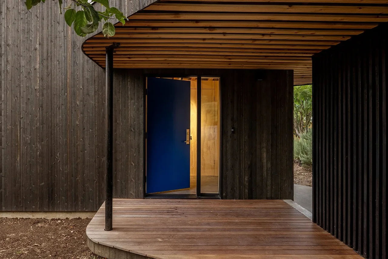 The entrance of House O featuring a blue front door and dark wood siding under a wood-paneled covered porch.