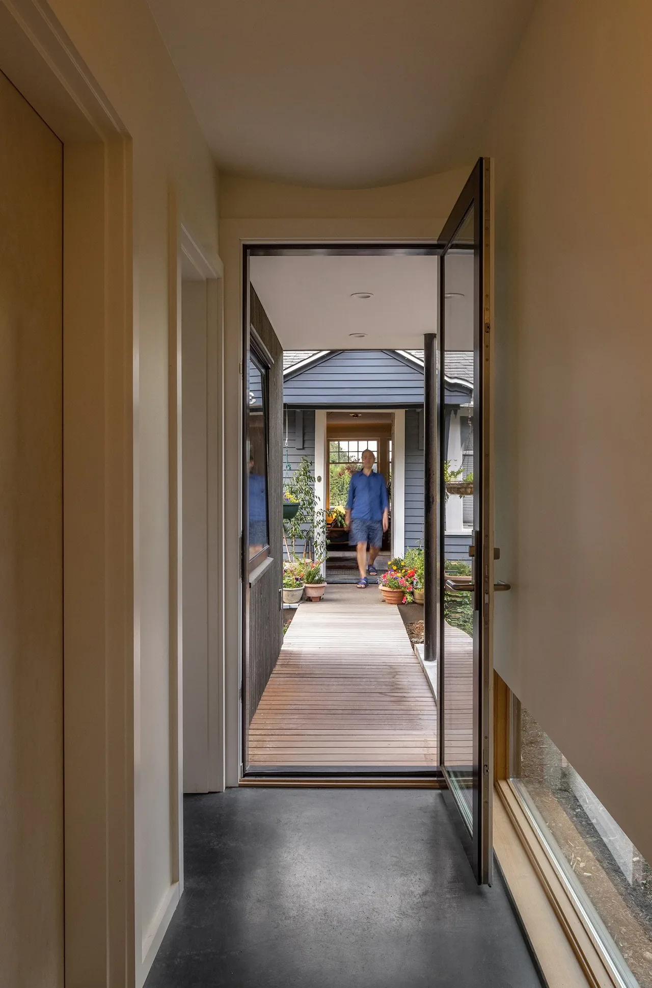 Hallway view through an open pivot glass door onto a covered wooden walkway with potted plants and a man walking outside.