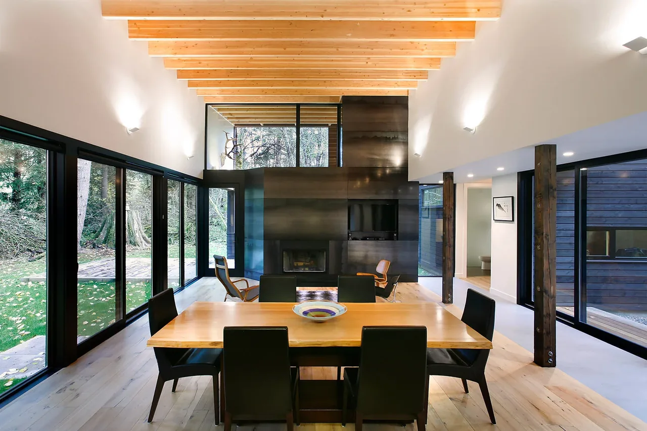 Dining area with exposed wood ceiling beams and a metal fireplace wall in a modern courtyard house on a river.