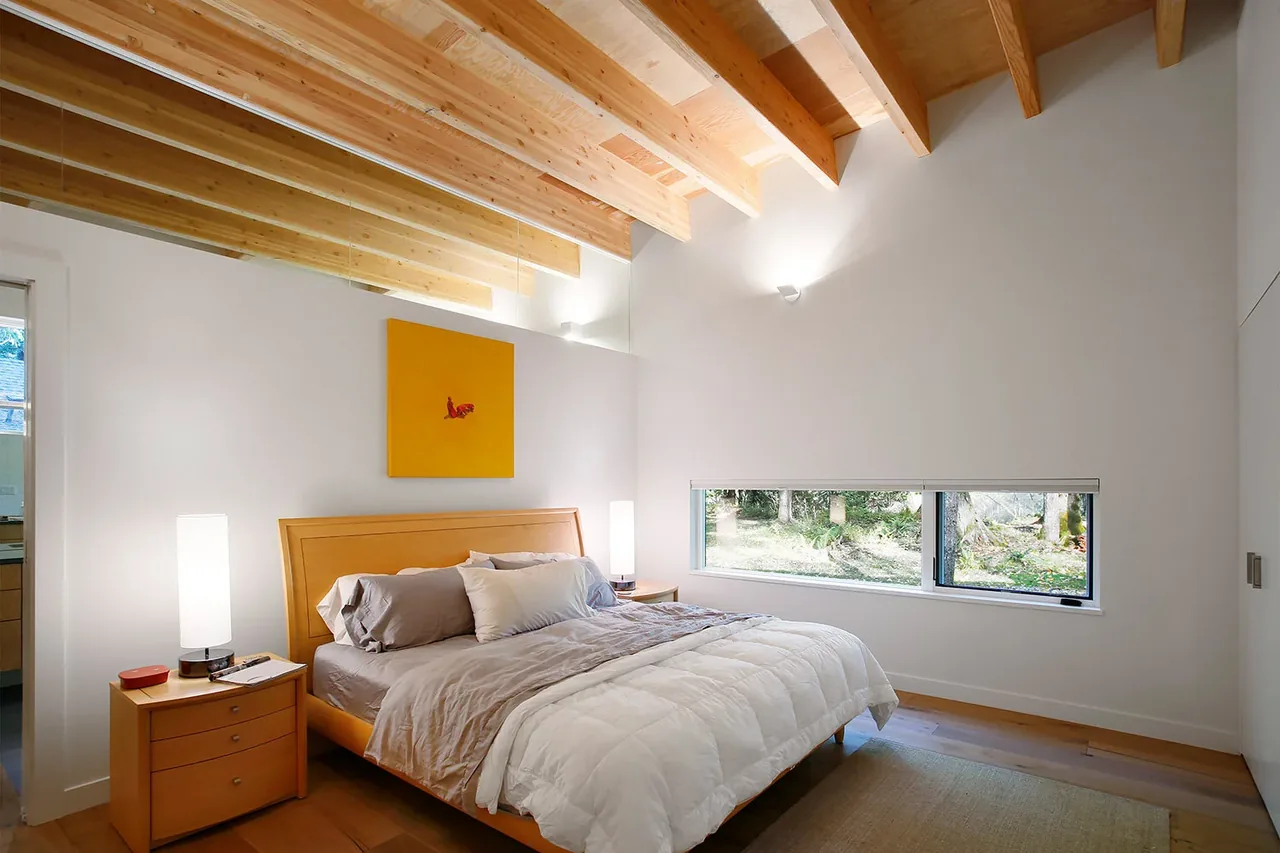 A bedroom featuring exposed wooden ceiling beams, a horizontal window, and simple contemporary furnishings in neutral tones.