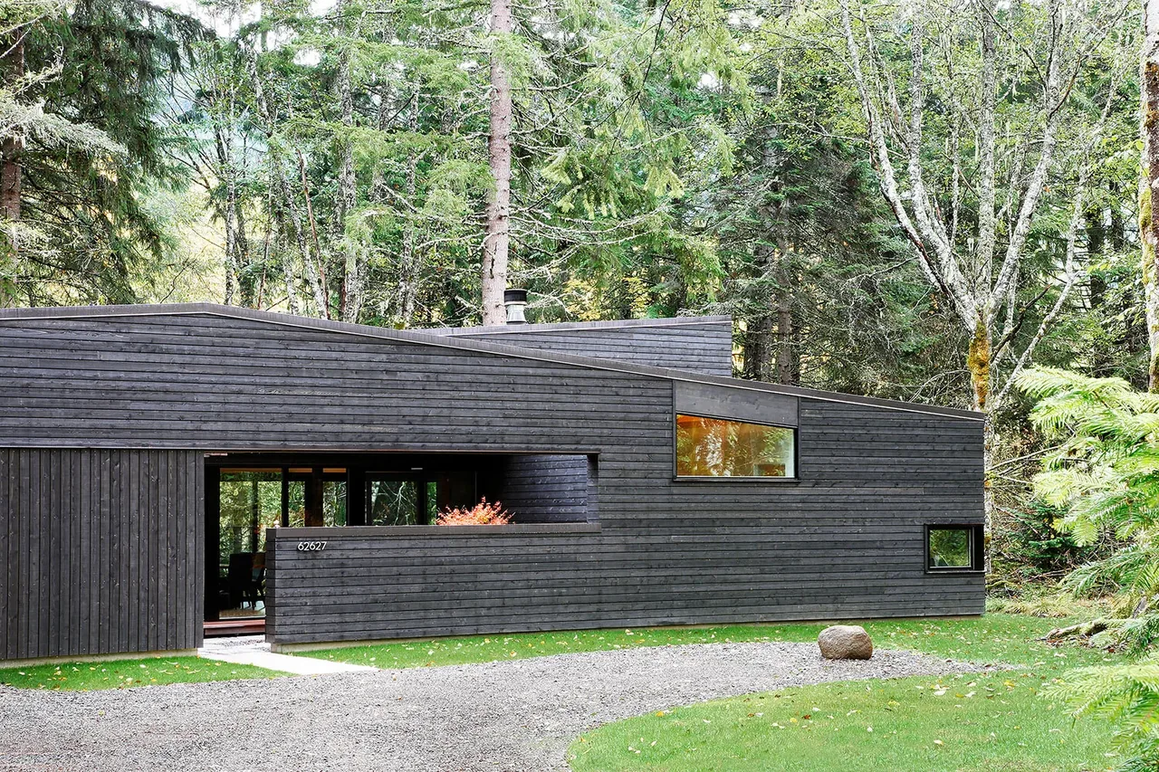 The Courtyard House on a River features dark wood siding and multiple flat roof planes set in a wooded landscape.