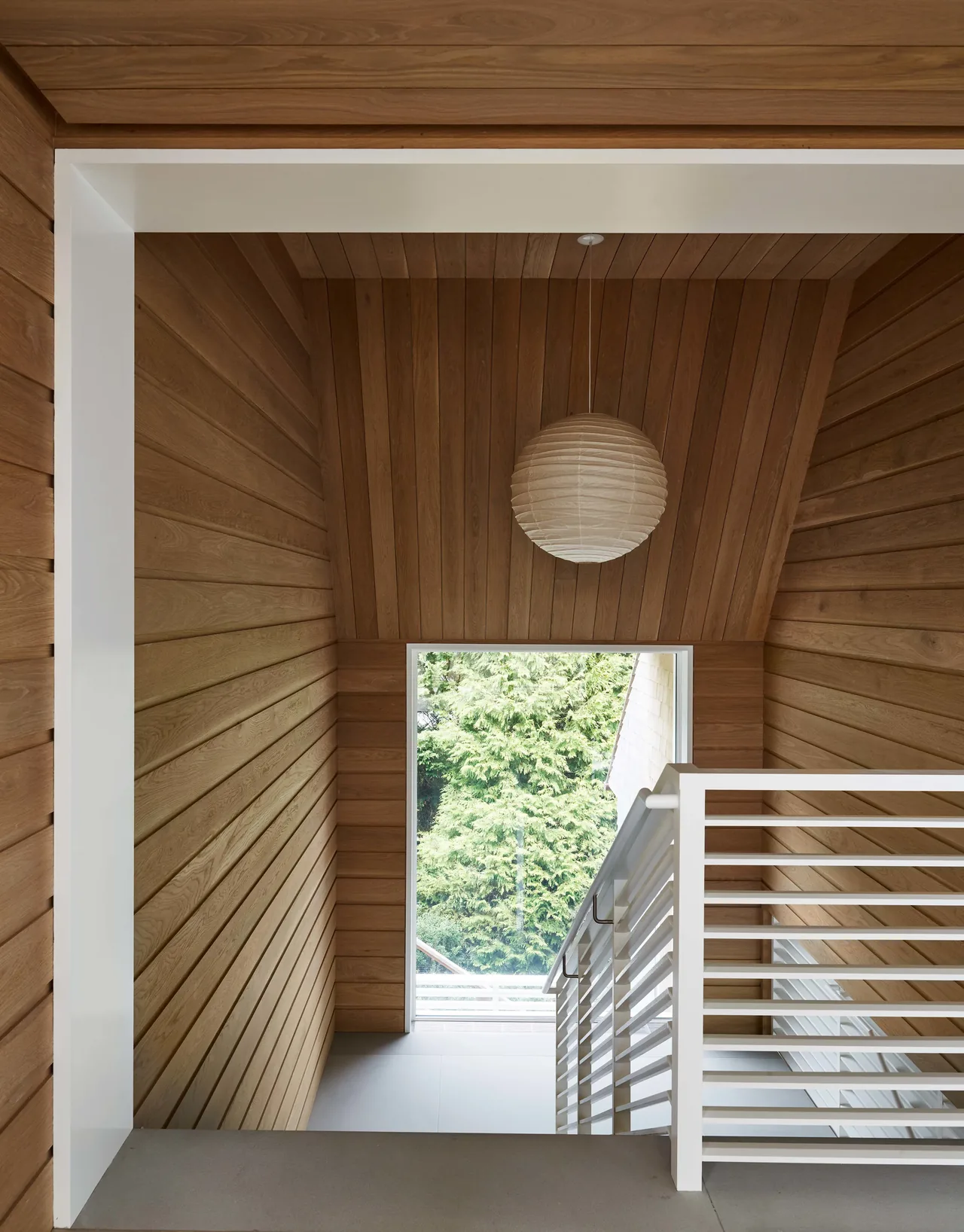 Stairwell clad in vertical wood panels with white metal railings and a spherical hanging light near a large window.