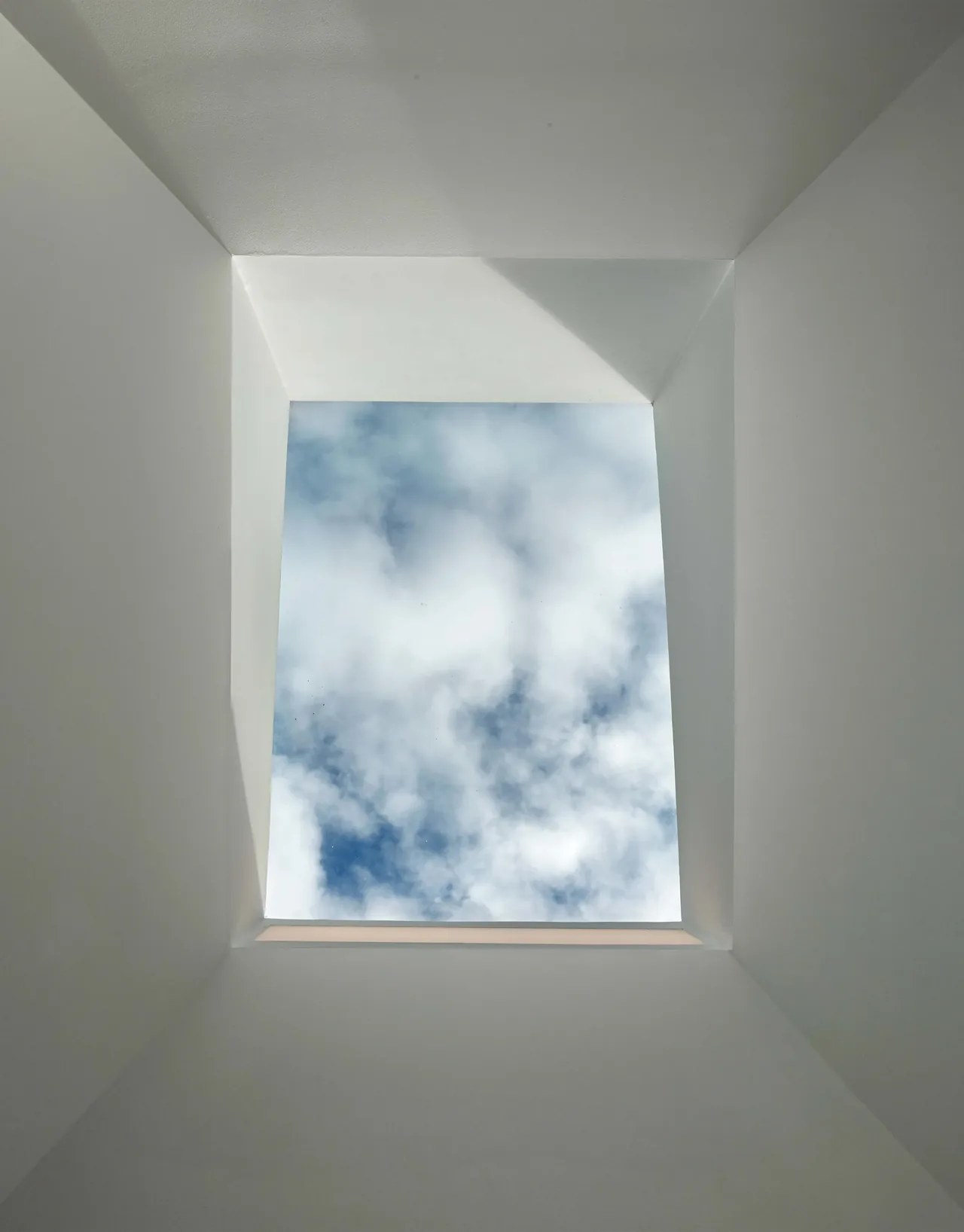A square skylight with white plaster walls showcasing a partly cloudy sky above.