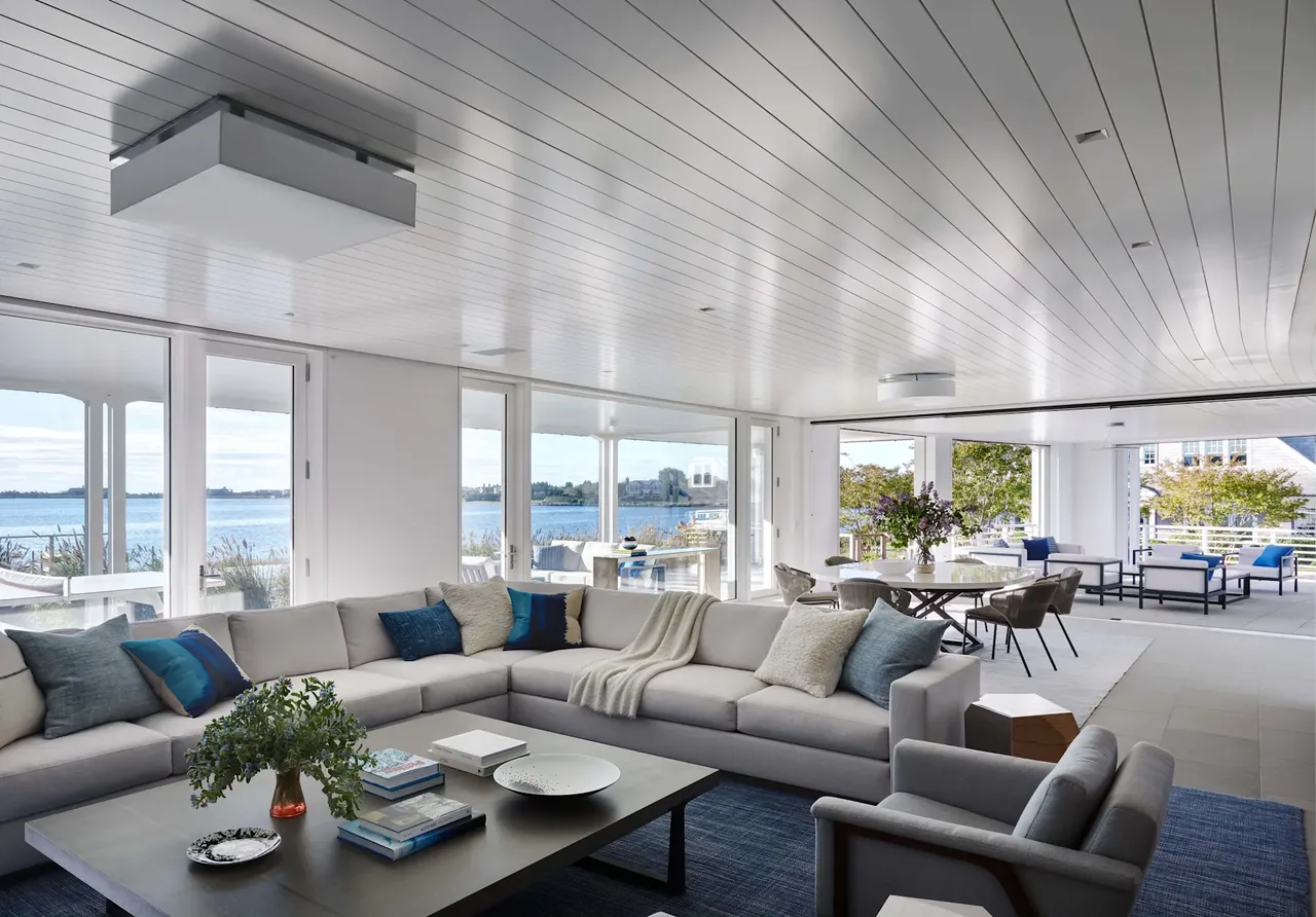Living room features a large sectional with abundant pillows and expansive glass doors revealing a waterfront view and connected outdoor seating area.