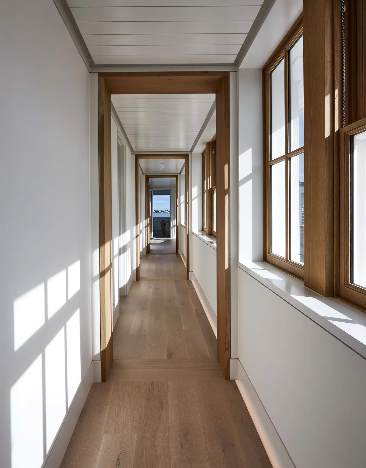 A bright hallway features wood-framed windows and flooring with a distant ocean view at the end of the corridor.