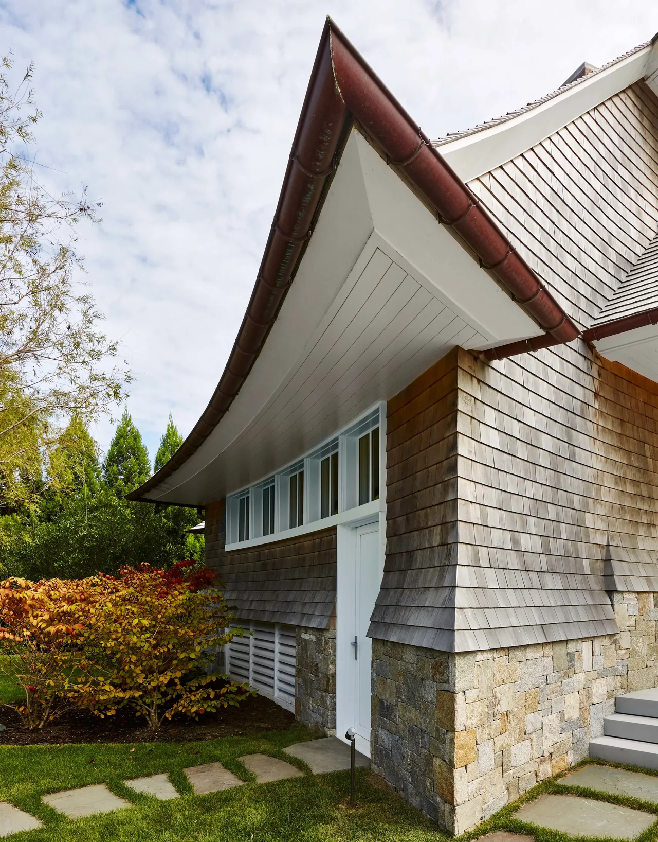 Water Mill House features curved roof eaves, wood shingle siding, and stacked stone lower walls in this exterior corner view.