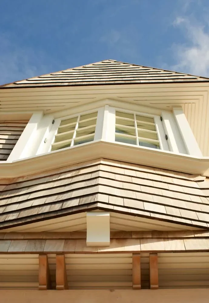 Exterior view of a shingle-sided home with a bay window and grid-patterned glass beneath a steep roof.