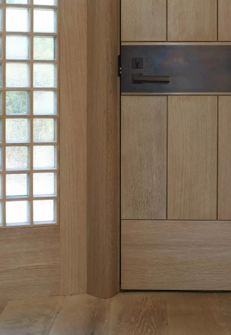Wood paneled door with vertical grooved panels and a metal handle beside a grid-patterned frosted glass window.