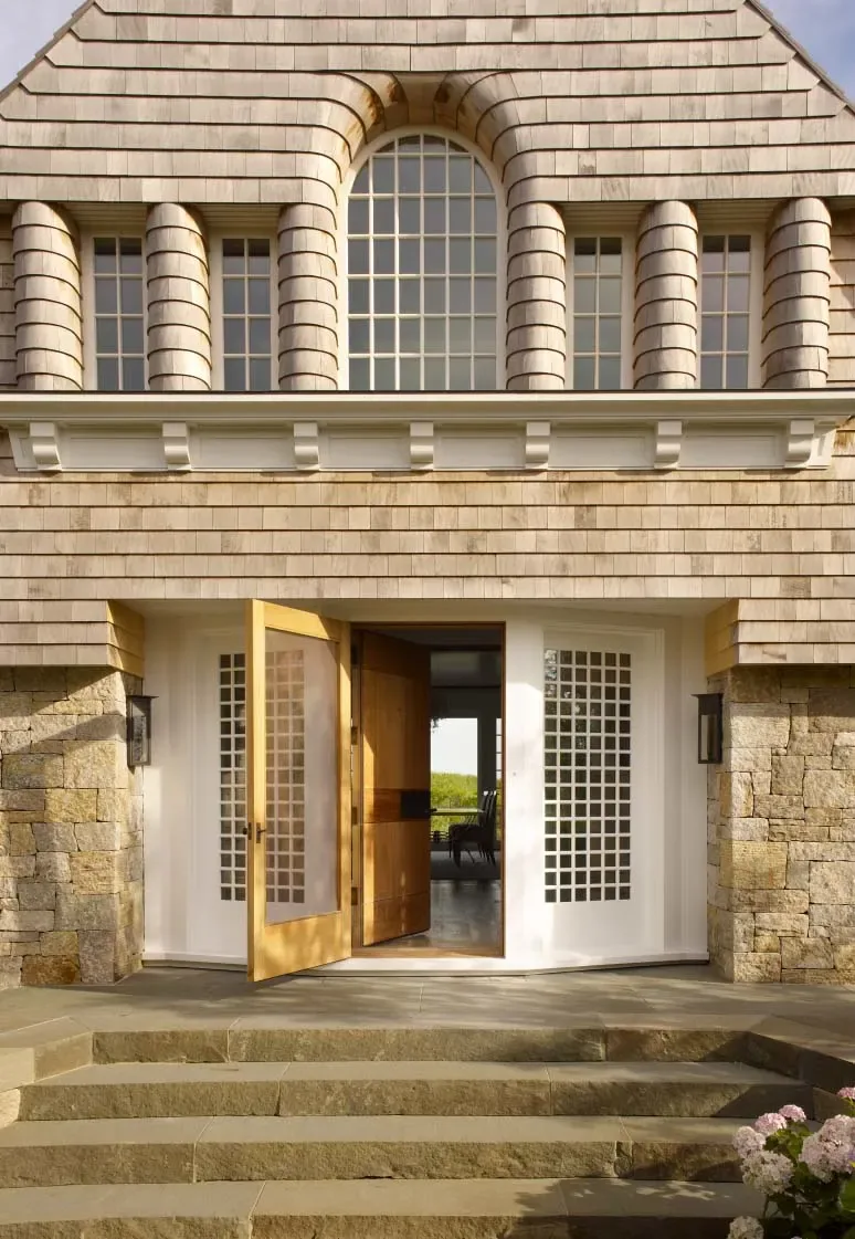 Front entry of a beach front house featuring wood shingles, stone steps, and a large wooden pivot door with grid windows.