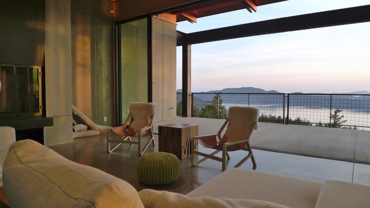 Living room with exposed beams and concrete flooring opens to an outdoor balcony with lake views at dusk.