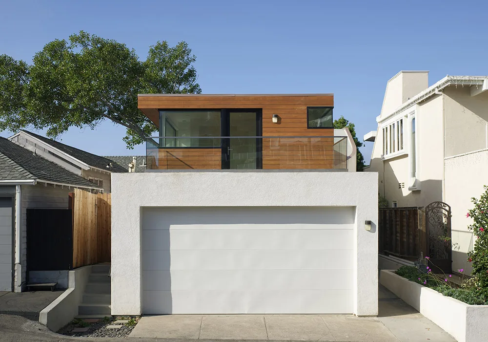 Modern two-story house with a white stucco garage base and upper wood-paneled level with glass railing under a clear sky.