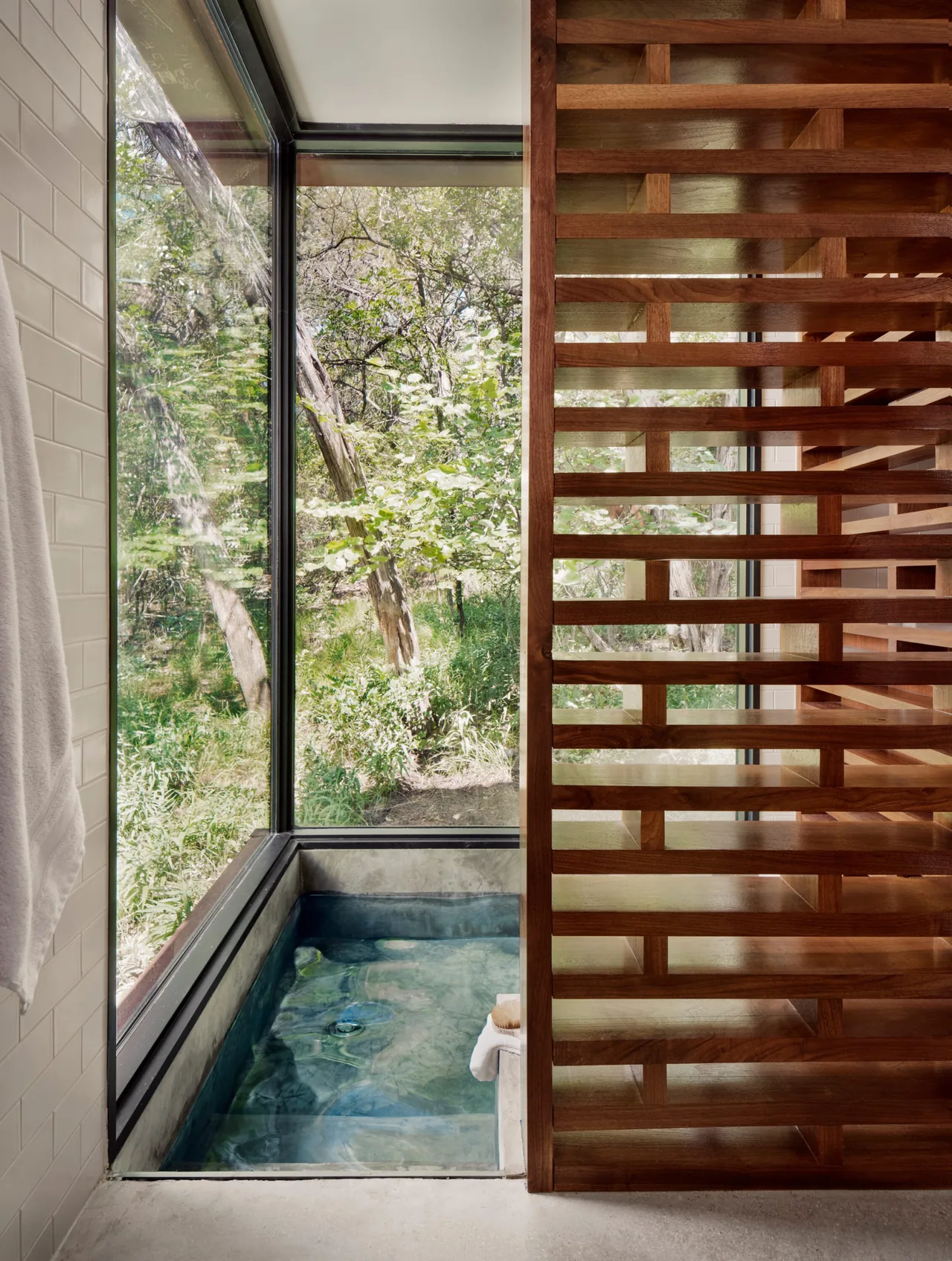A sunken bath beside a corner window overlooking a wooded landscape, partially separated by a wooden slatted partition.