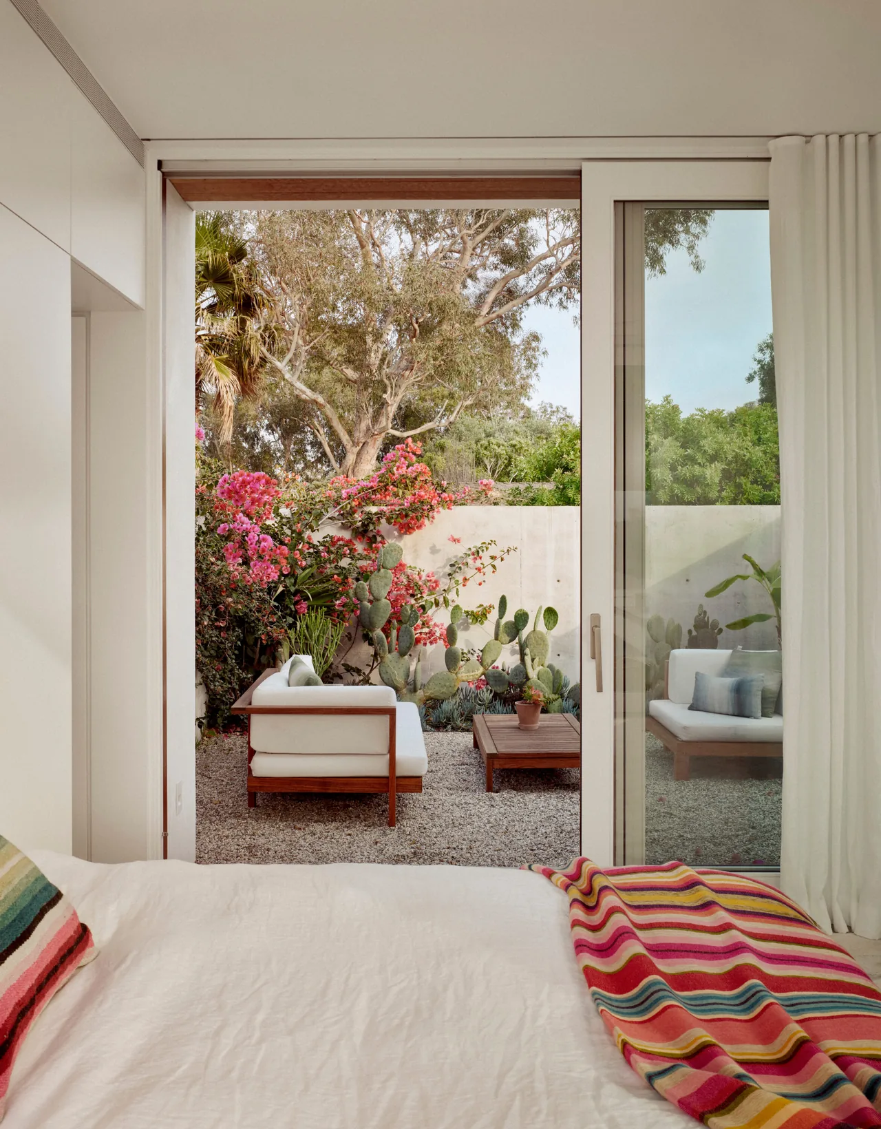 Bedroom opening to a small patio with wooden outdoor furniture and lush greenery including pink flowers and cactus.