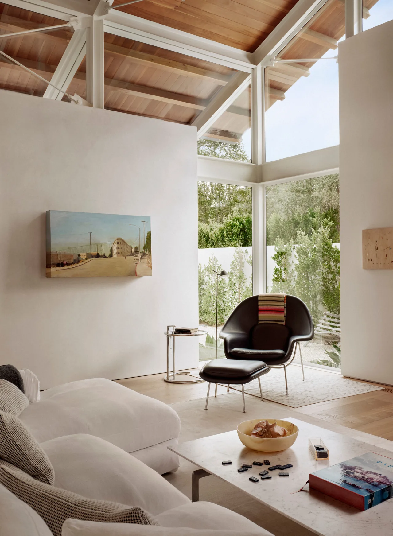 A living room in Point Dume Residence features vaulted wood beam ceilings and large corner windows with a black armchair and white sofa.