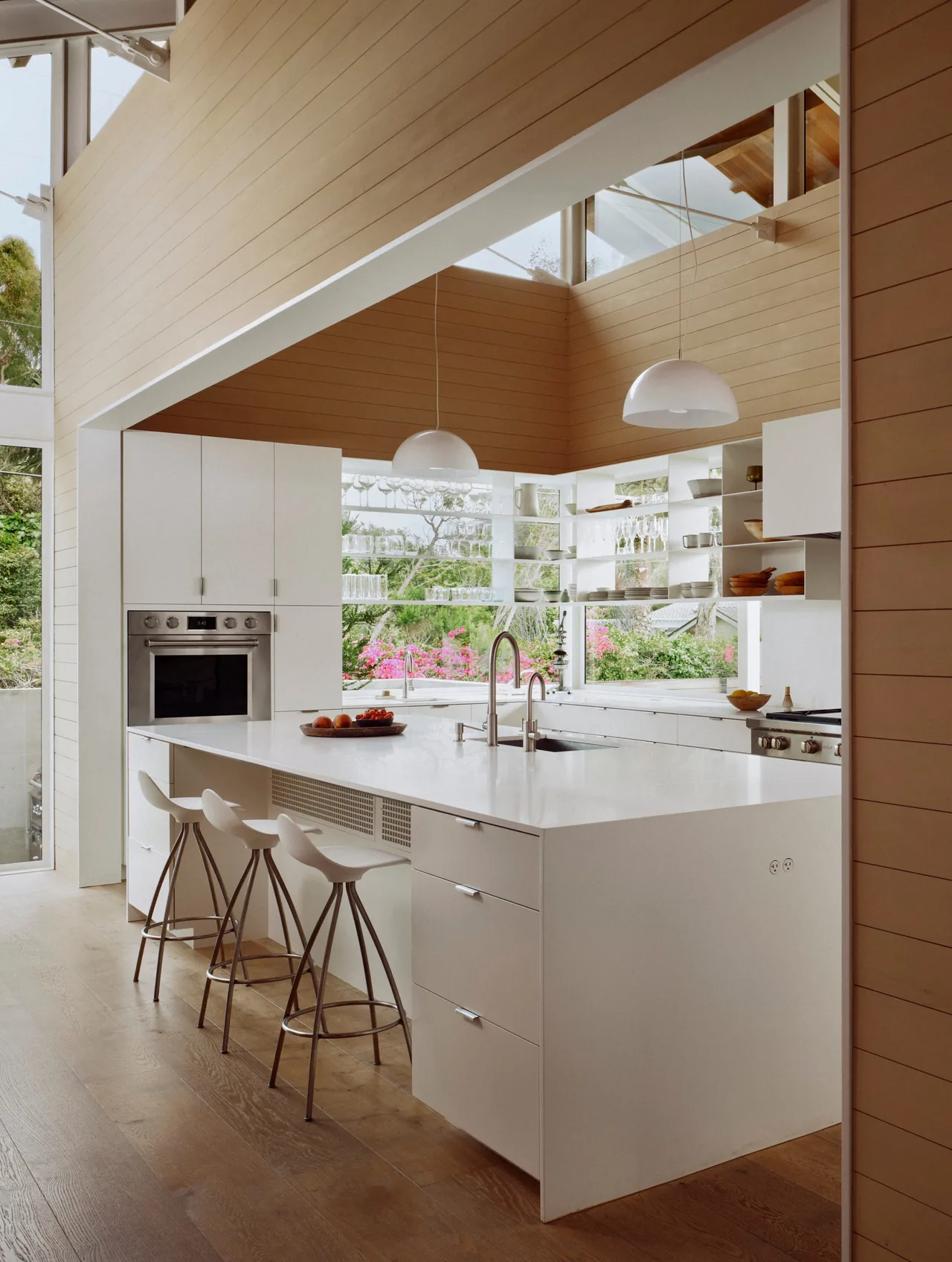 The Point Dume Residence kitchen features a large white island, wood beam ceiling, and expansive windows with garden views.