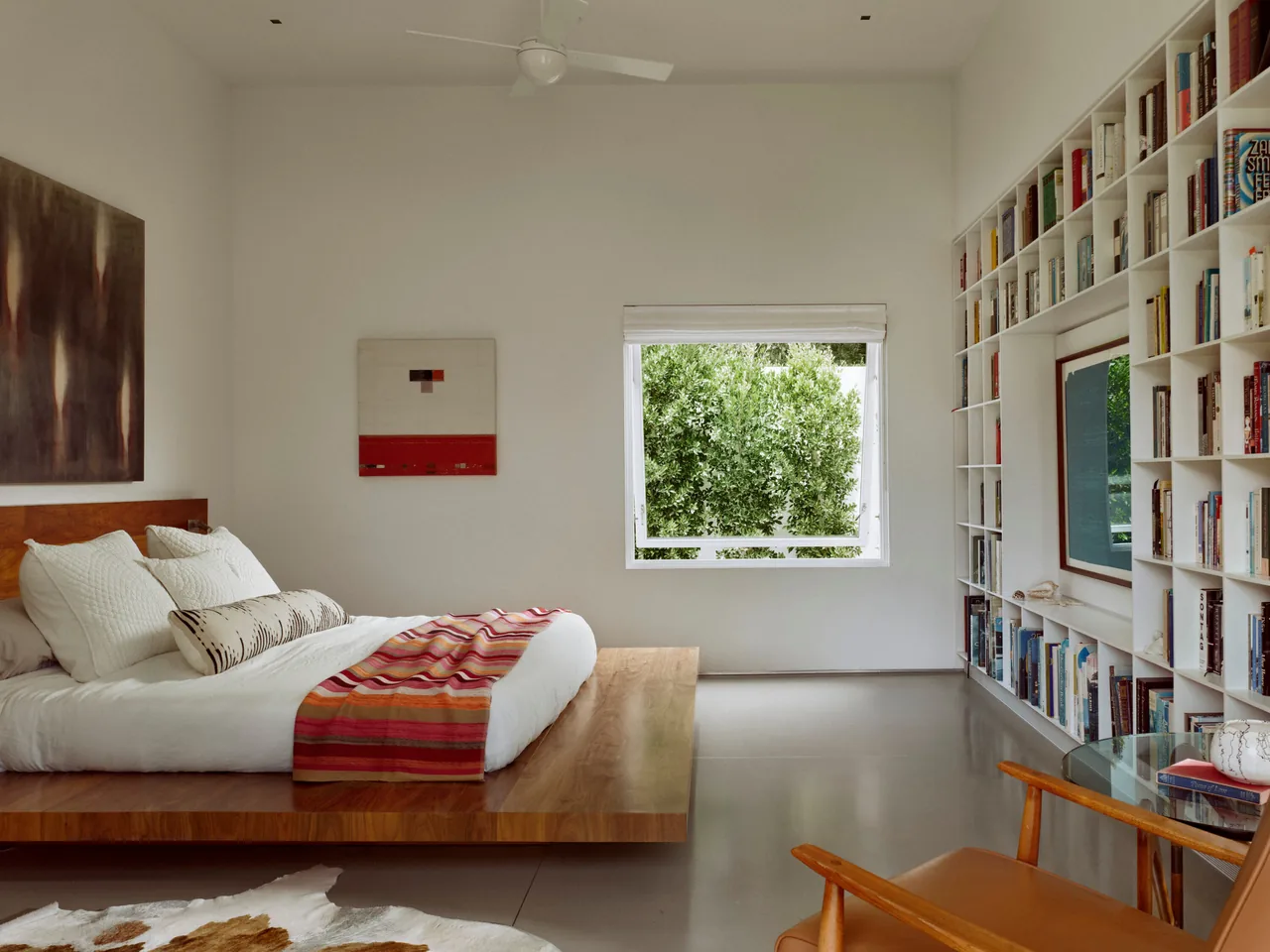 Minimalist bedroom featuring a wooden platform bed and a built-in bookcase wall with a view of trees through a window.