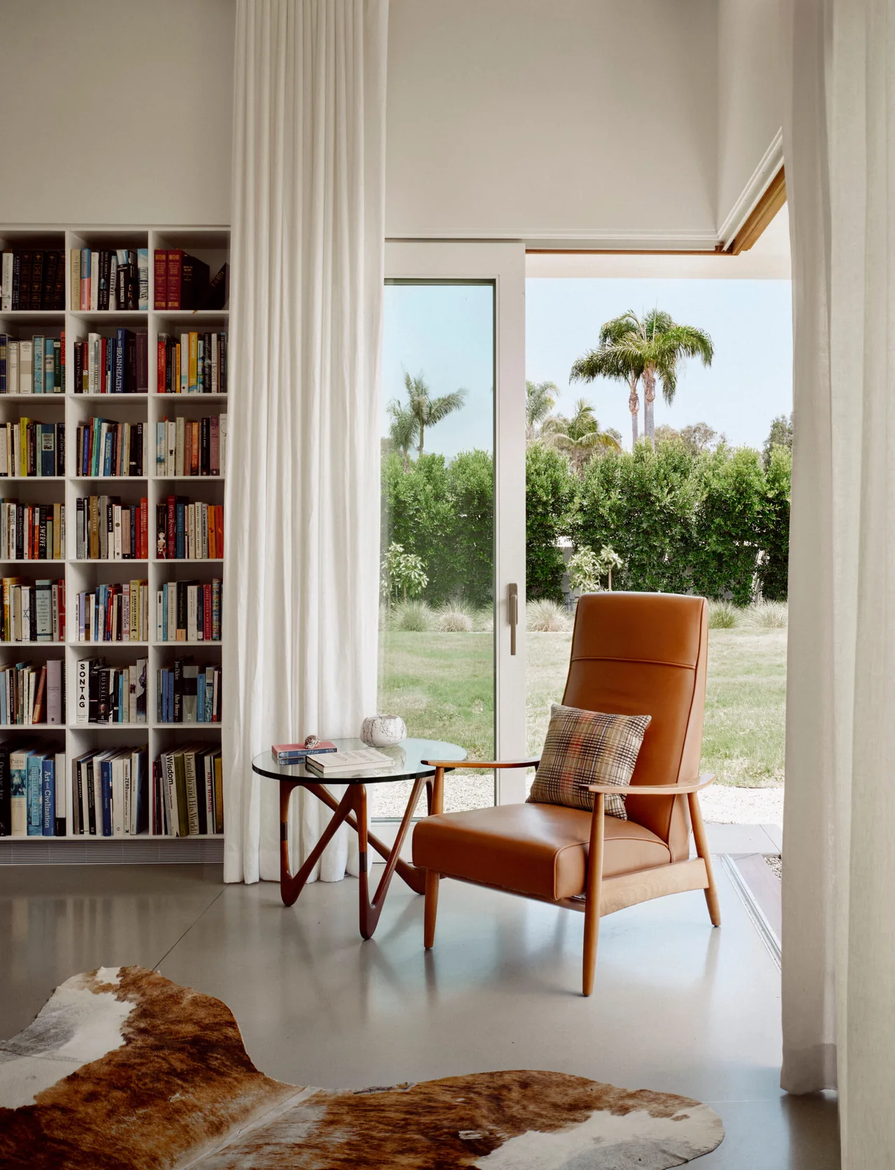 Seating area with a leather chair and glass-top wooden side table next to a sliding glass door and a built-in bookcase.