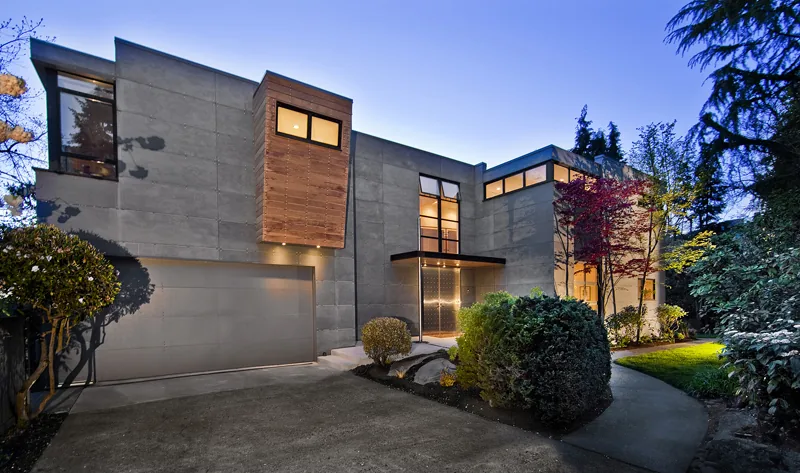 Modern Mercer Island home remodel with concrete exterior, wood accents, and large dark-framed windows at dusk.