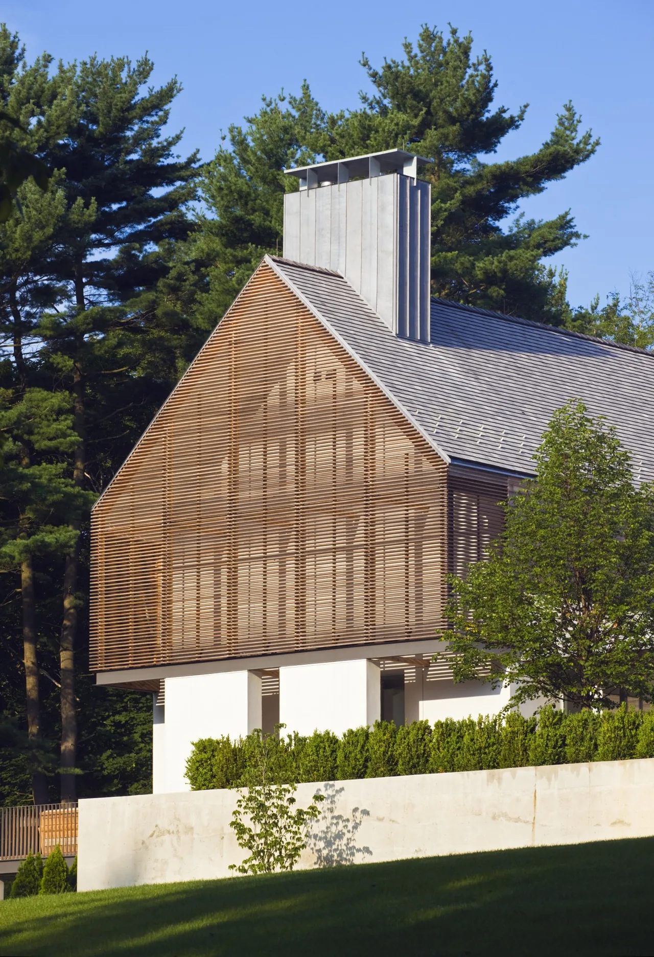 Modern house featuring a wooden slatted gable, white walls, and a metal chimney, surrounded by trees and landscaping.