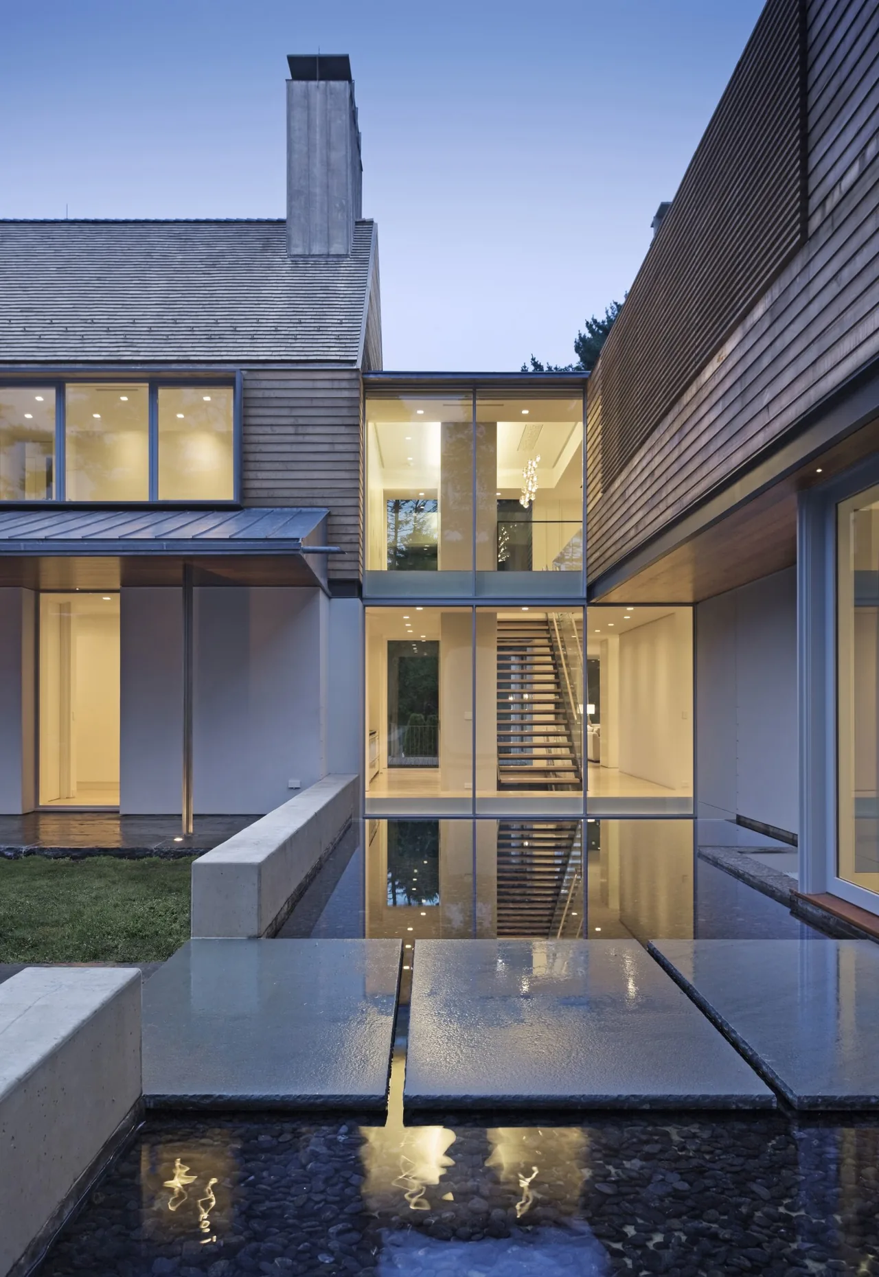 Exterior view of a modern house at dusk featuring large glass windows, a water feature, and a visible stairway inside.
